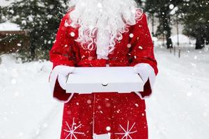 scatola della pizza bianca nelle mani di babbo natale in guanti bianchi, con la barba, in un mantello rosso. consegna di fast food di natale. promozione di capodanno. lavoro nei giorni festivi di ristorazione. copia spazio, mock up foto