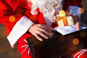 Babbo Natale con un laptop e un regalo in mano. ordina i regali di natale e capodanno attraverso il negozio online. prenotazione dei servizi di un animatore per le vacanze. foto