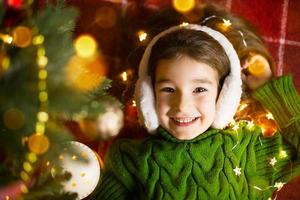 una ragazza felice con le cuffie di pelliccia giace vicino all'albero di natale in ghirlande e luci in un maglione caldo su un'accogliente coperta. natale e capodanno, vigilia delle vacanze, atmosfera magica foto