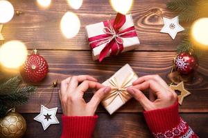 le mani delle donne in maglione lavorato a maglia con ornamento tengono una scatola con un regalo di natale nei palmi delle mani sul tavolo tra l'arredamento. capodanno, confezionamento e preparazione di regali, rami di abete, decorazione foto