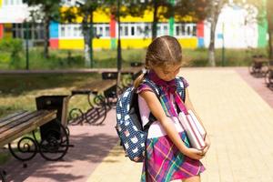 ragazza con uno zaino e una pila di libri vicino alla scuola. Tornato a scuola, il bambino è stanco, libri di testo pesanti. istruzione, classi della scuola primaria, inizio anno scolastico, 1 settembre foto