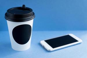 tazza usa e getta in carta kraft bianca per caffè con coperchio in plastica nera e telefono cellulare bianco su sfondo blu. foto