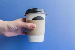 tazza usa e getta in carta kraft bianca per caffè con coperchio in plastica nera in una mano femminile. caffè per andare su sfondo blu. foto