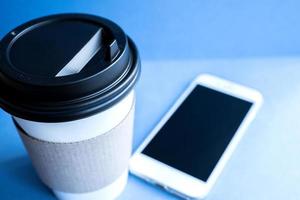tazza usa e getta in carta kraft bianca per caffè con coperchio in plastica nera e telefono cellulare bianco su sfondo blu. foto