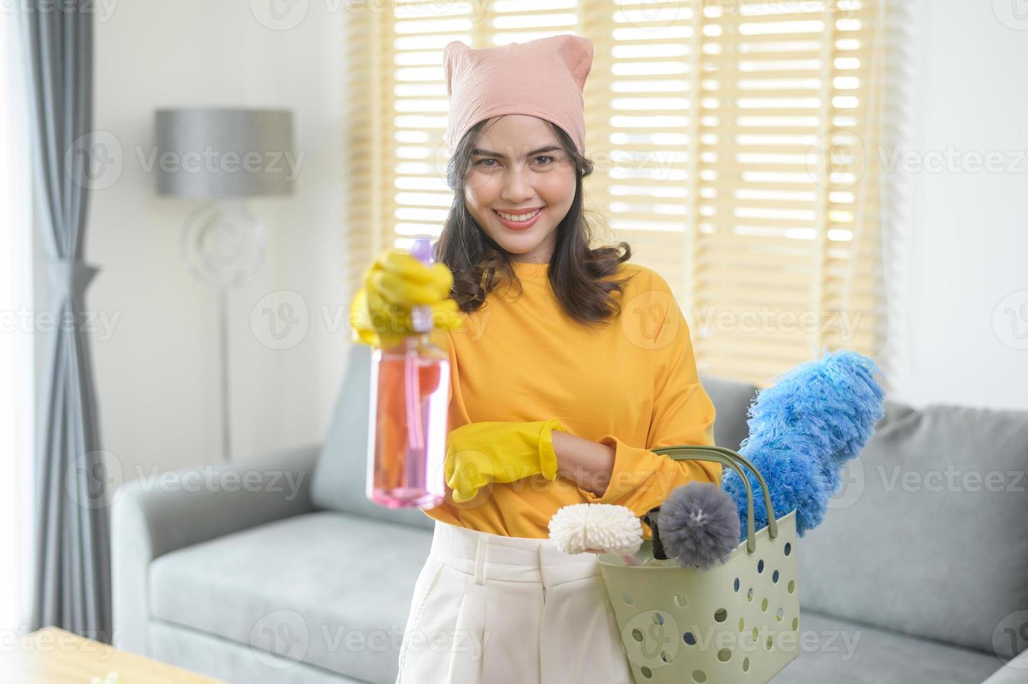 jovem mulher feliz usando luvas amarelas e segurando uma cesta de material de limpeza na sala de estar. foto