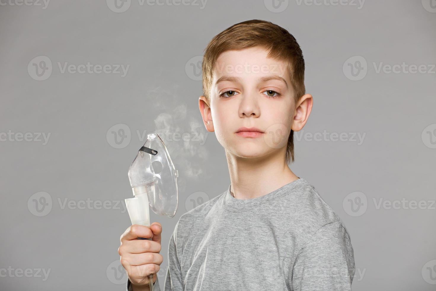 menino infeliz segurando a máscara de inalação, liberando fumaça no fundo cinza foto