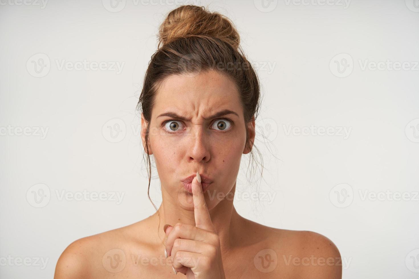 foto interna de mulher muito mal-humorada, levantando o dedo indicador aos lábios, pedindo para manter o silêncio, franzindo a testa e olhando seriamente para a câmera