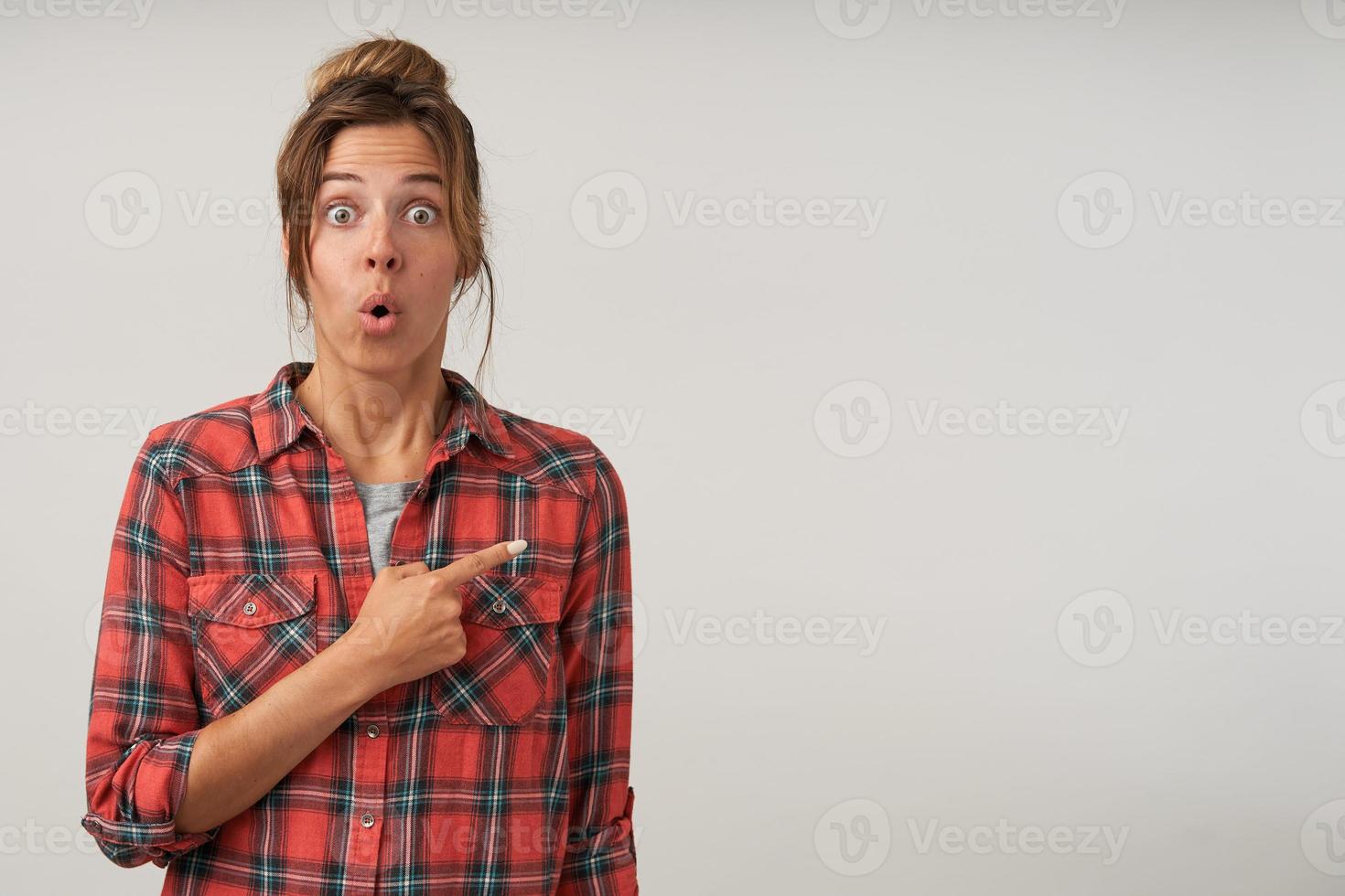 foto de estúdio de jovem mulher espantada em roupas casuais posando sobre fundo branco, apontando com o dedo indicador de lado, olhando para a câmera com os olhos bem abertos