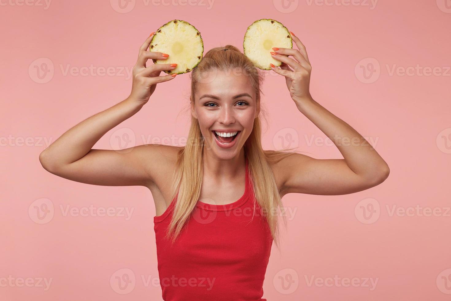 foto de estúdio de encantadora jovem com longos cabelos loiros posando sobre fundo rosa na camisa vermelha, tirando sarro com anéis de abacaxi fresco, sorrindo alegremente para a câmera