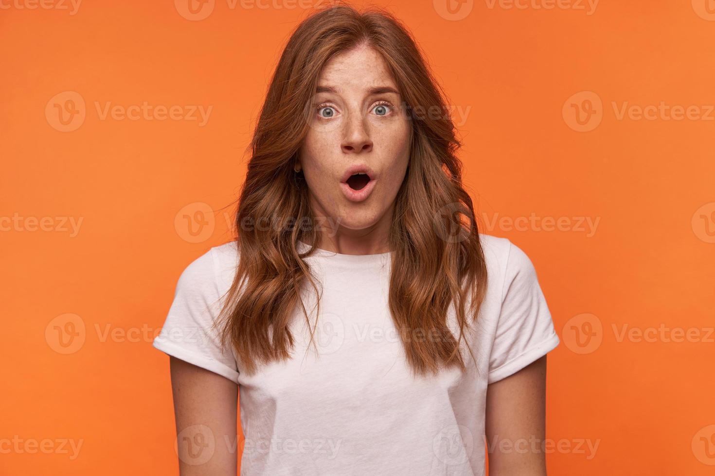 foto de estúdio de chocada mulher jovem com cabelo ruivo encaracolado em pé sobre fundo laranja, olhando surpresa para a câmera com a boca aberta