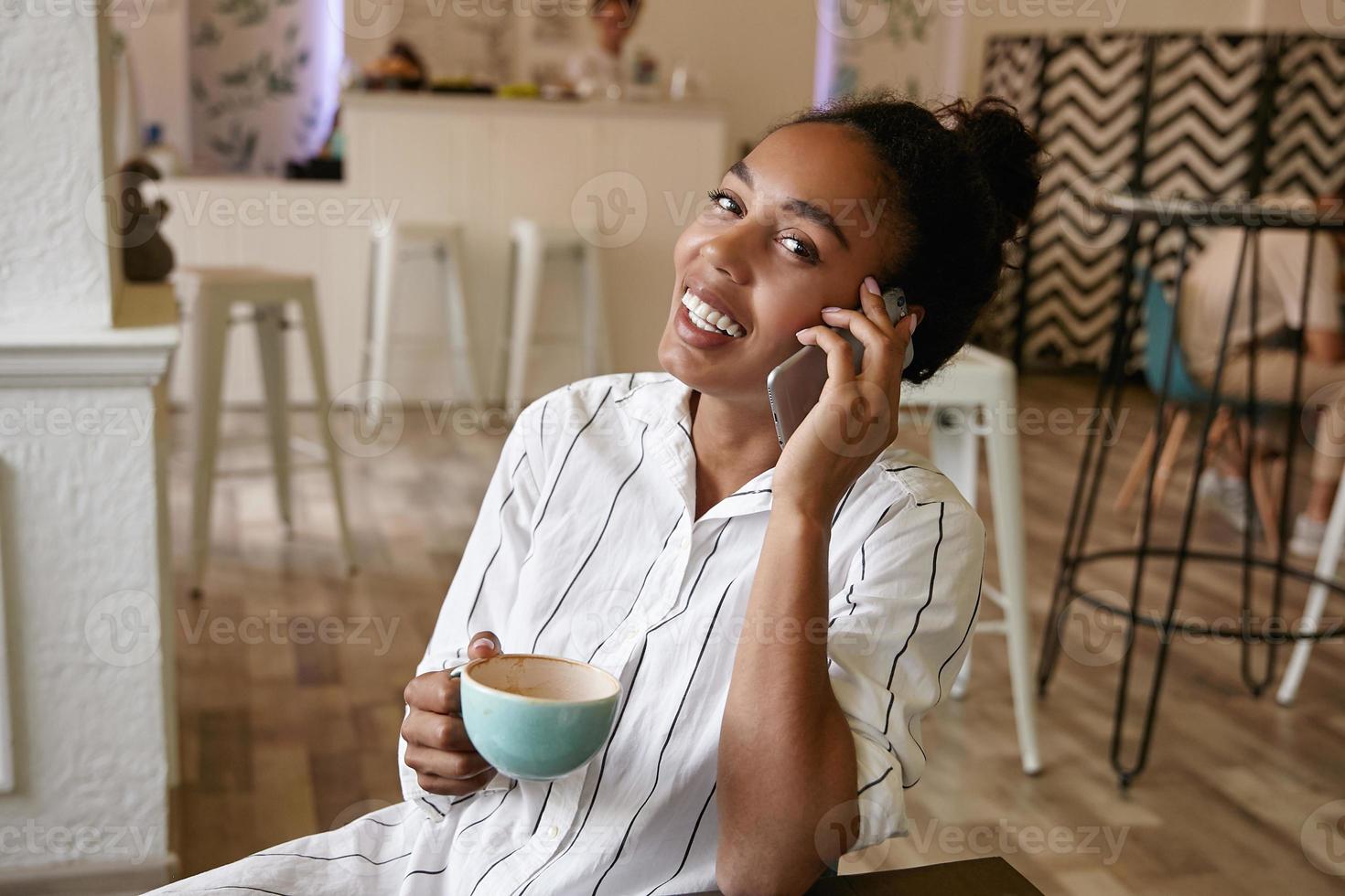 mulher de pele escura atraente feliz com penteado de coque falando no telefone com um amigo enquanto bebe café no café, olhando para a câmera alegremente foto