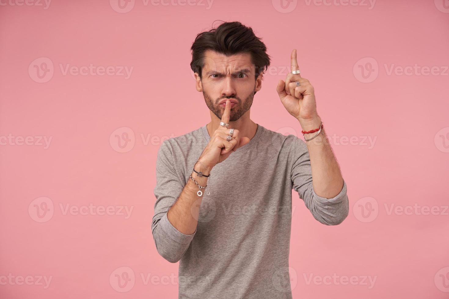 homem bonito sério com corte de cabelo na moda, vestindo suéter cinza, posando sobre fundo rosa com os dedos levantados em gesto de silêncio, chamando para manter o silêncio, franzindo a testa e franzindo os lábios foto