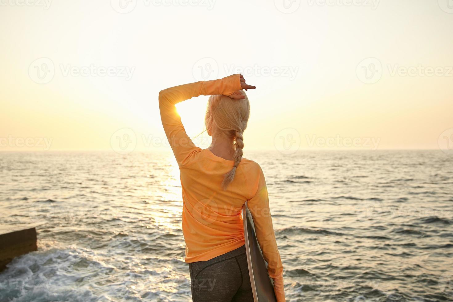 vista traseira ao ar livre da jovem mulher loira magro em roupas casuais, olhando para o nascer do sol e mantendo o braço na cabeça, posando à beira-mar com prancha de equilíbrio na mão foto