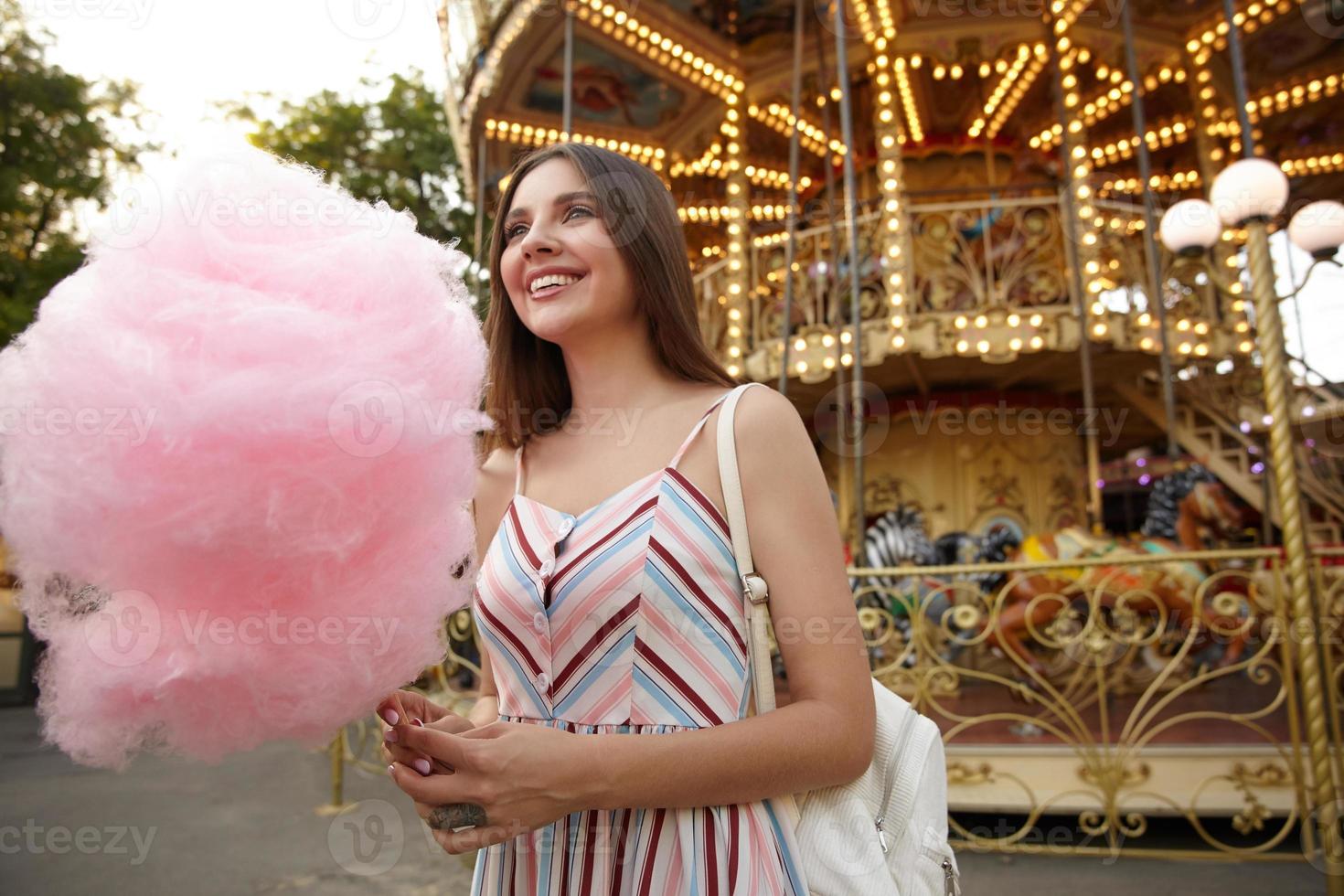 tiro ao ar livre da morena de cabelos compridos jovem atraente usando vestido de verão, posando sobre carrossel em dia quente, segurando algodão doce na vara, olhando de lado com um sorriso sincero foto