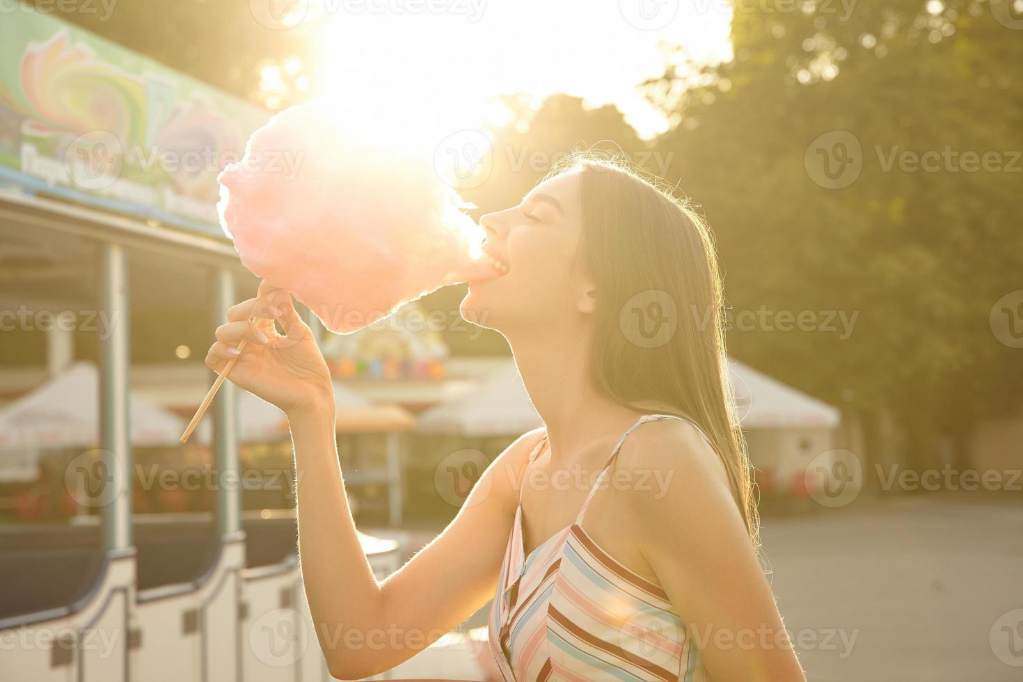 foto ensolarada ao ar livre de feliz senhora de cabelos compridos em vestido romântico posando sobre parque de diversões em dia quente de verão, comendo algodão doce rosa com grande prazer