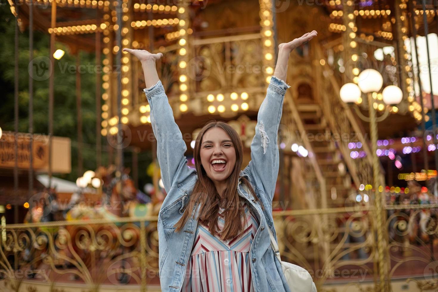 feliz jovem morena com cabelo comprido em pé sobre carrossel no parque de atrações, levantando as mãos alegremente com a boca aberta, conceito de emoções positivas foto