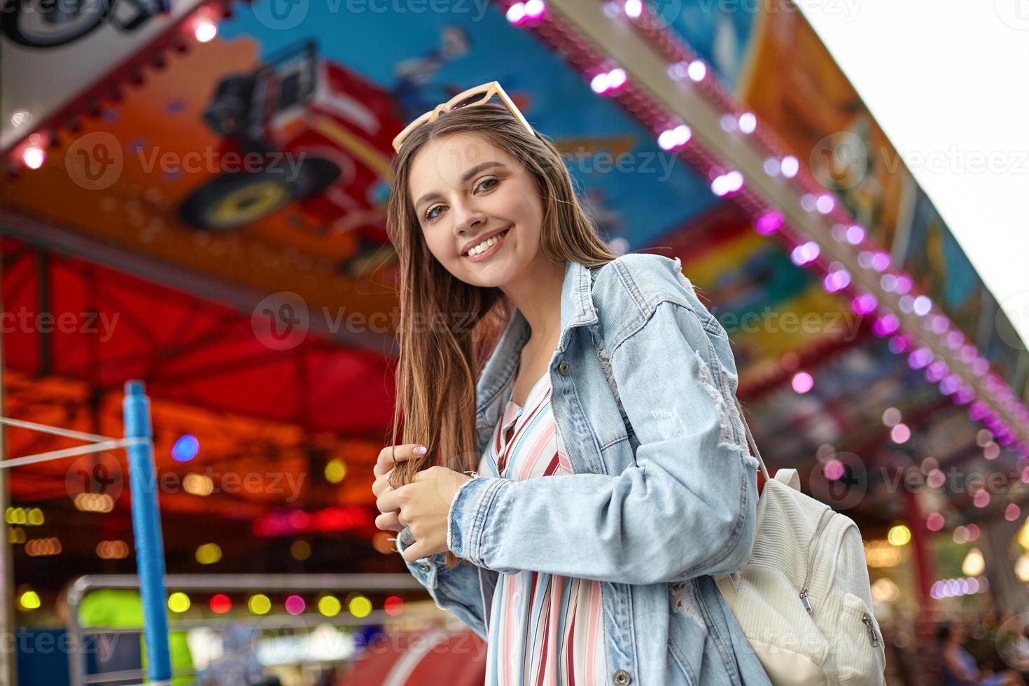 foto ao ar livre de uma jovem bonita positiva com longos cabelos castanhos, olhando para a câmera com um sorriso sincero, vestindo mochila branca, casaco jeans da moda e vestido leve de verão