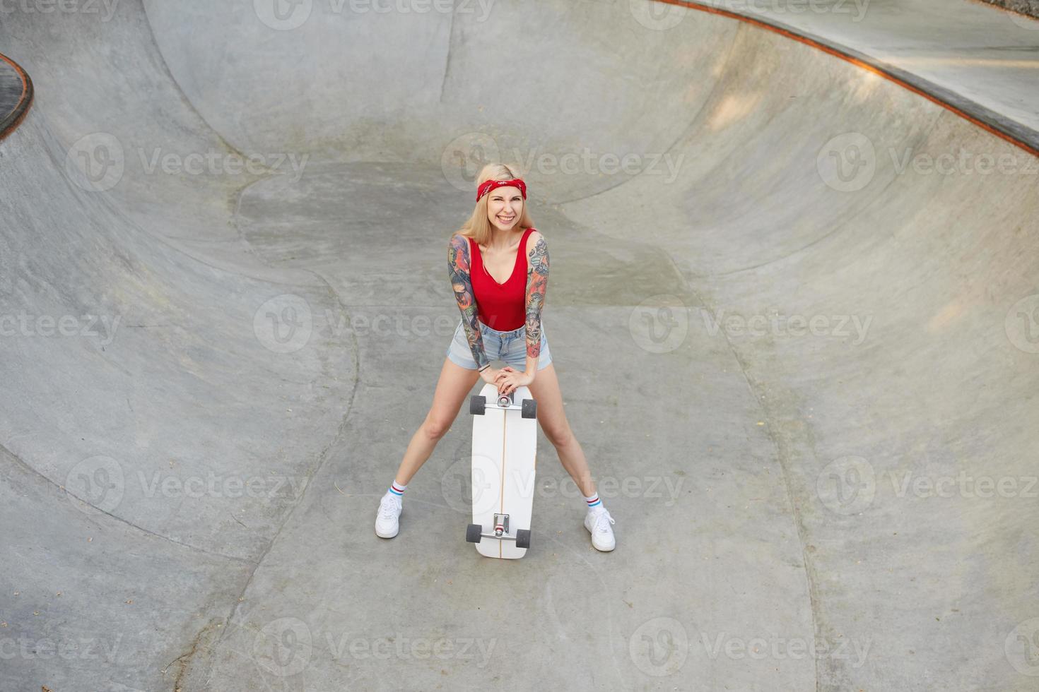 alegre mulher loira tatuada de cabelos compridos posando sobre skate park em dia quente e brilhante, vestindo shorts jeans e blusa vermelha, mantendo a prancha nas mãos e sorrindo alegremente foto