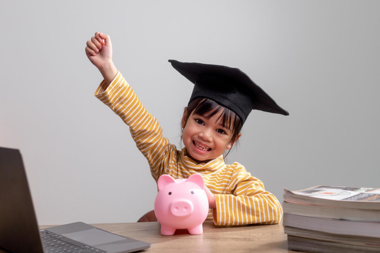menina asiática vestindo um boné de formatura com um cofrinho rosa, economizando dinheiro, investindo no futuro, foto