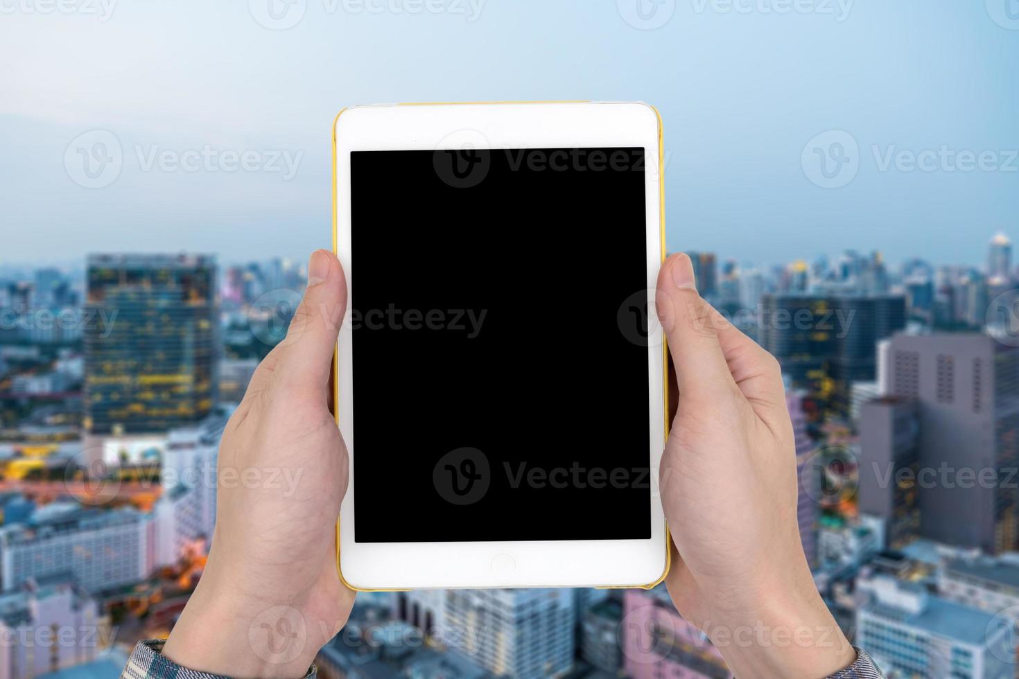 mãos masculinas segurando um gadget de computador de toque de tablet branco com tela preta em branco de toque no fundo da paisagem da cidade turva. foto