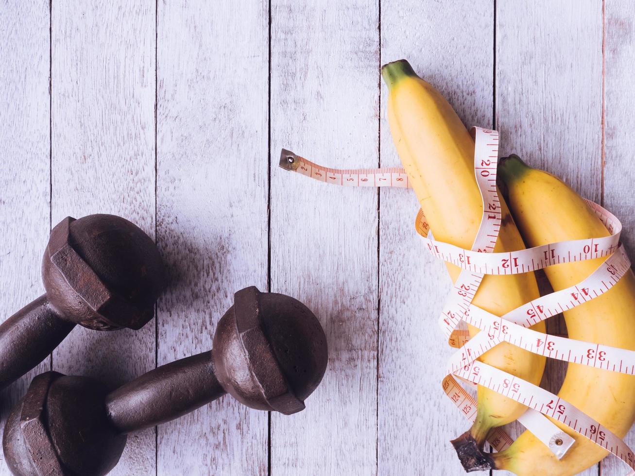 bananas com fita métrica e halteres de ferro no fundo da mesa de madeira. conceito de treino e dieta foto