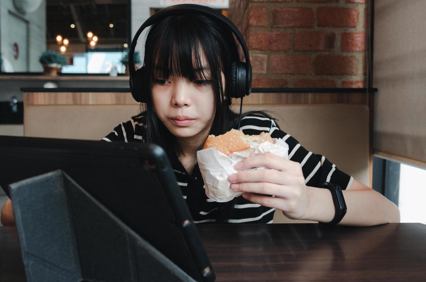 garota comendo hambúrguer assista seu tablet e use fones de ouvido para ouvir música ou usar as mídias sociais. foto