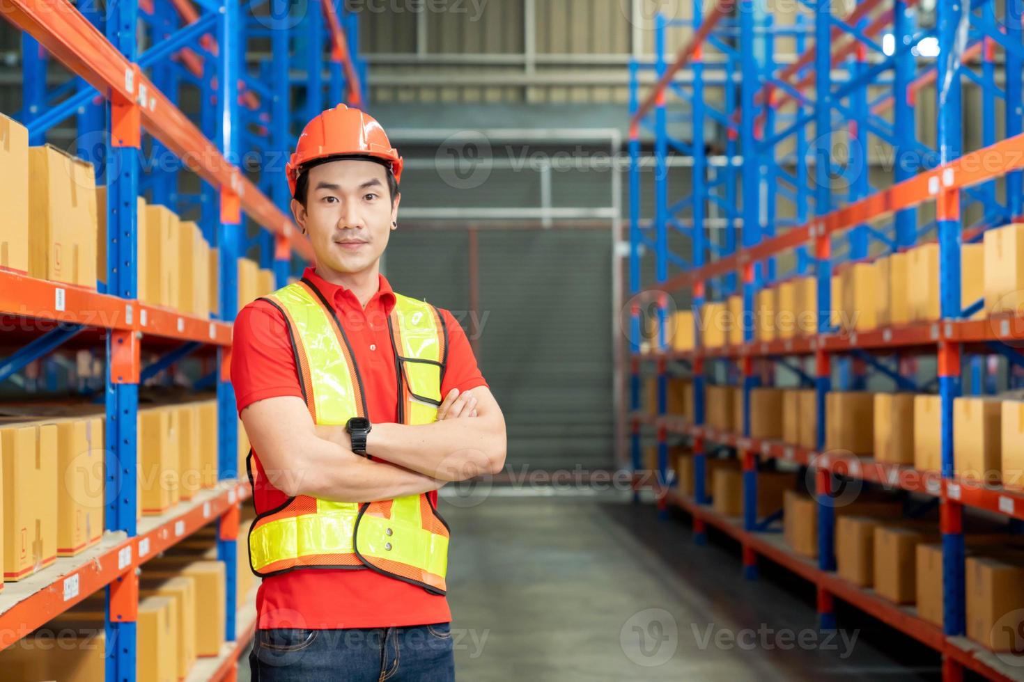 trabalhador de unidade de fabricação bem sucedido em pé no centro de distribuição do armazém com as mãos postas com capacete e uniforme sorrindo foto