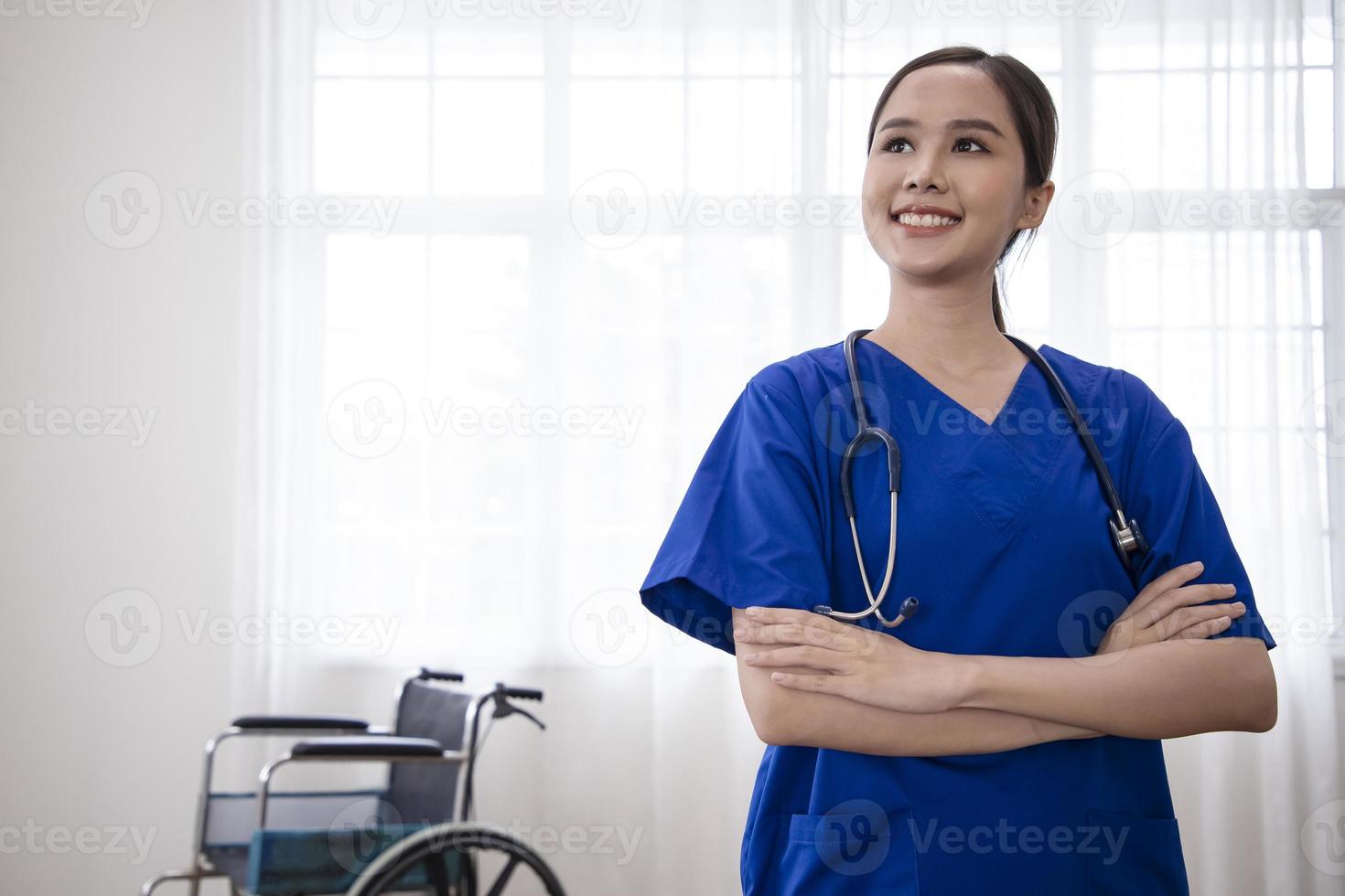 jovem enfermeira asiática sorrindo alegremente com uma mão no quadril e atitude confiante, positiva, orgulhosa e amigável contra a cortina branca com cadeira de rodas no fundo foto