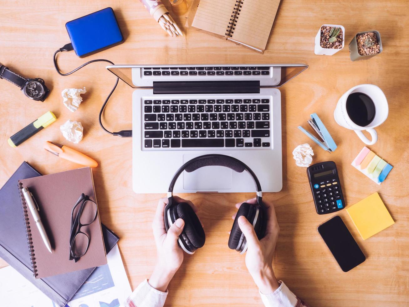 vista superior das mãos masculinas segurando fones de ouvido, laptop com material de escritório na mesa de madeira. foto