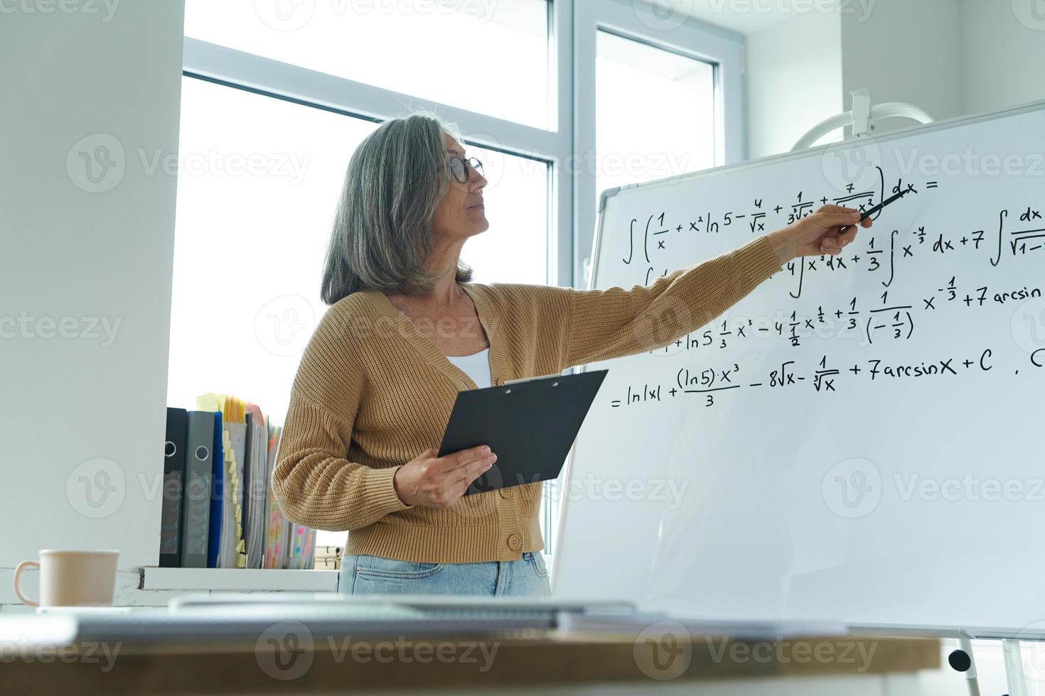 mulher sênior confiante ensinando matemática enquanto aponta o quadro branco na sala de aula foto