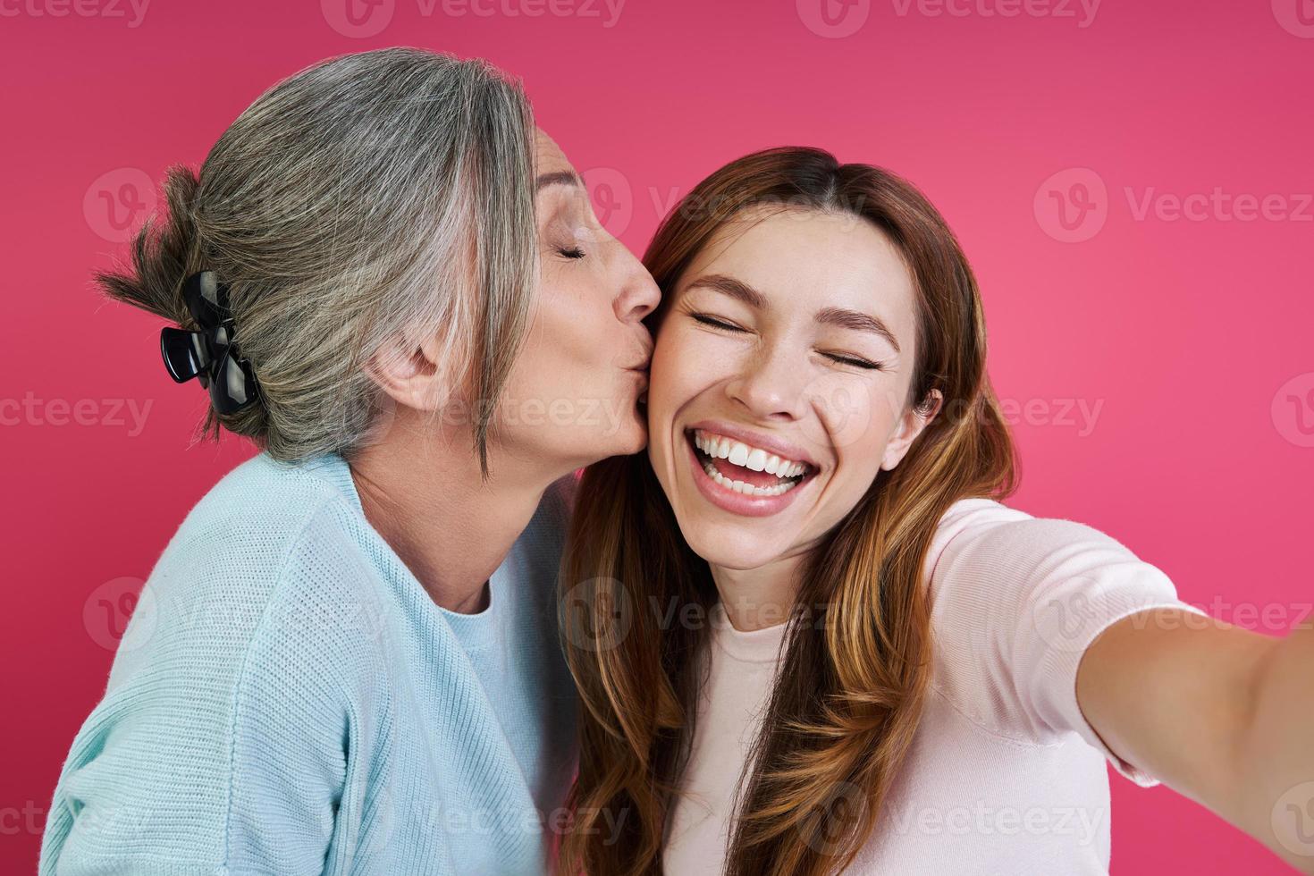 mãe sênior beijando sua filha alegre enquanto fazia selfie contra fundo rosa foto