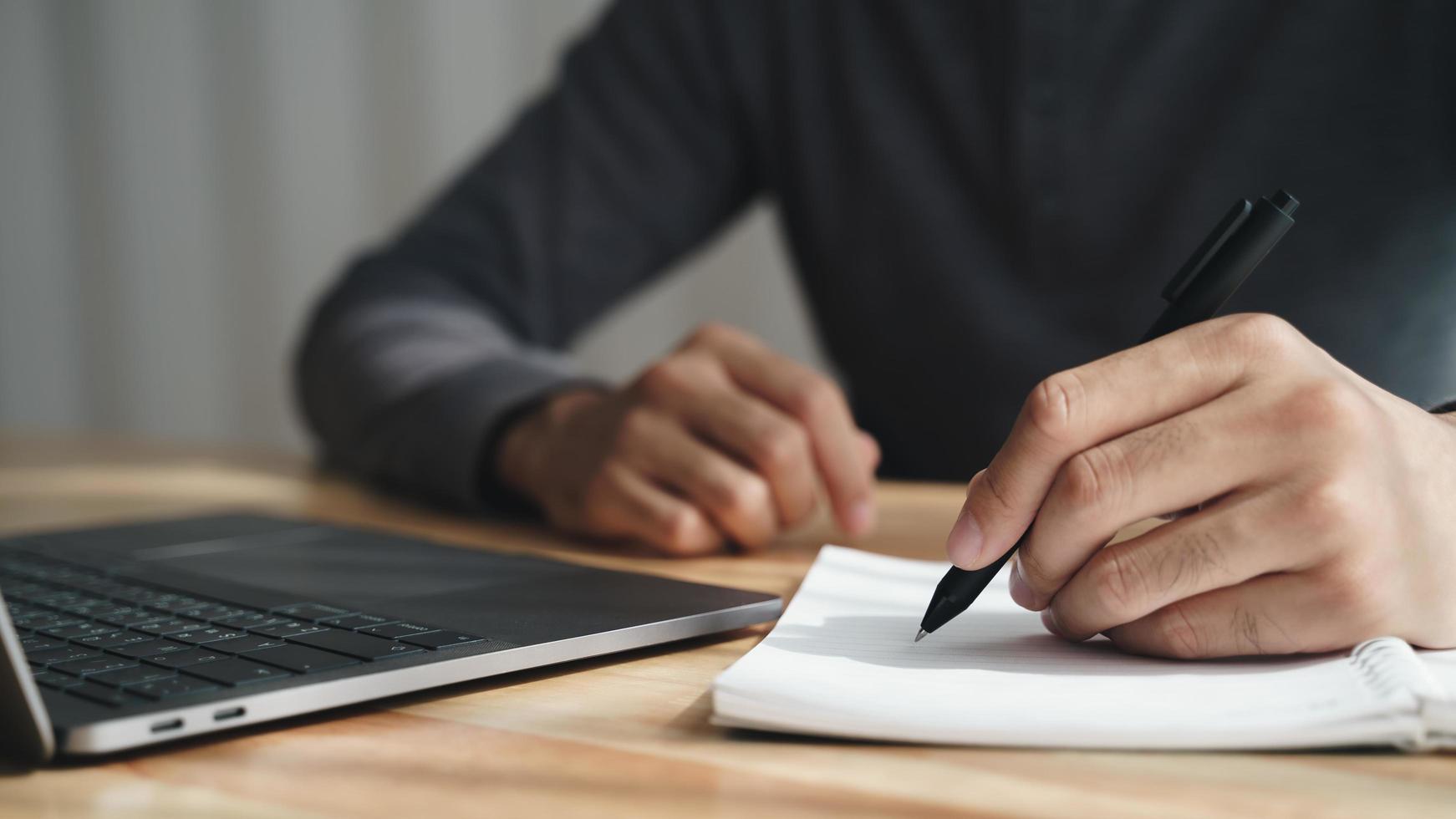 homem canhoto escreve em um caderno em cima da mesa com computador portátil foto