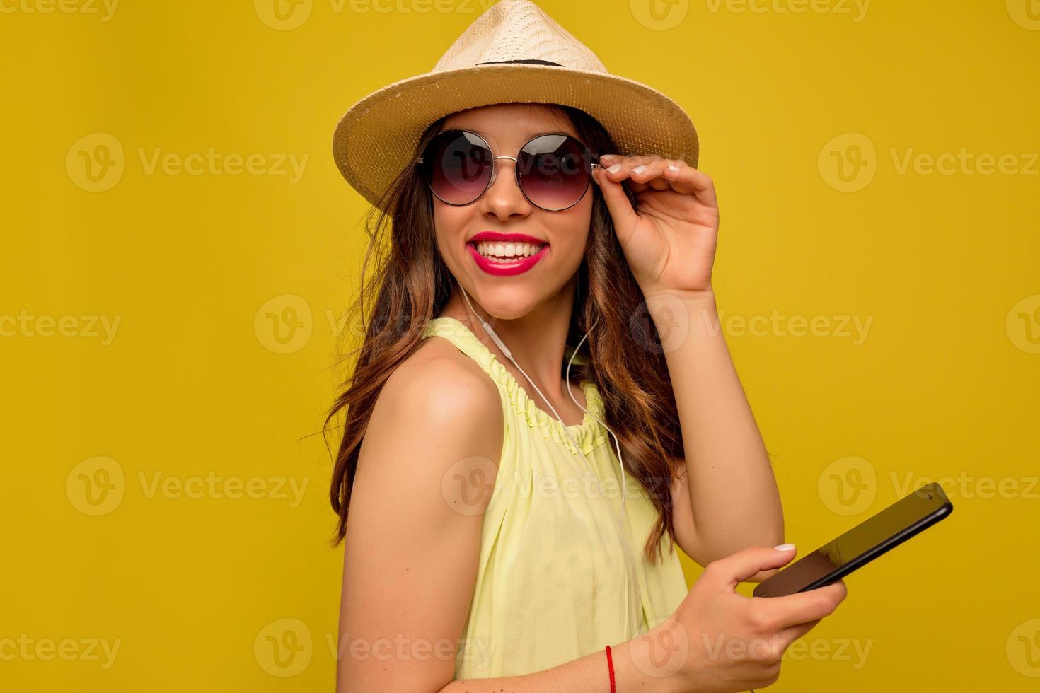 um retrato de uma menina feliz brilhante no chapéu de verão e óculos de sol usando smartphone posando no estúdio sobre o fundo amarelo. foto