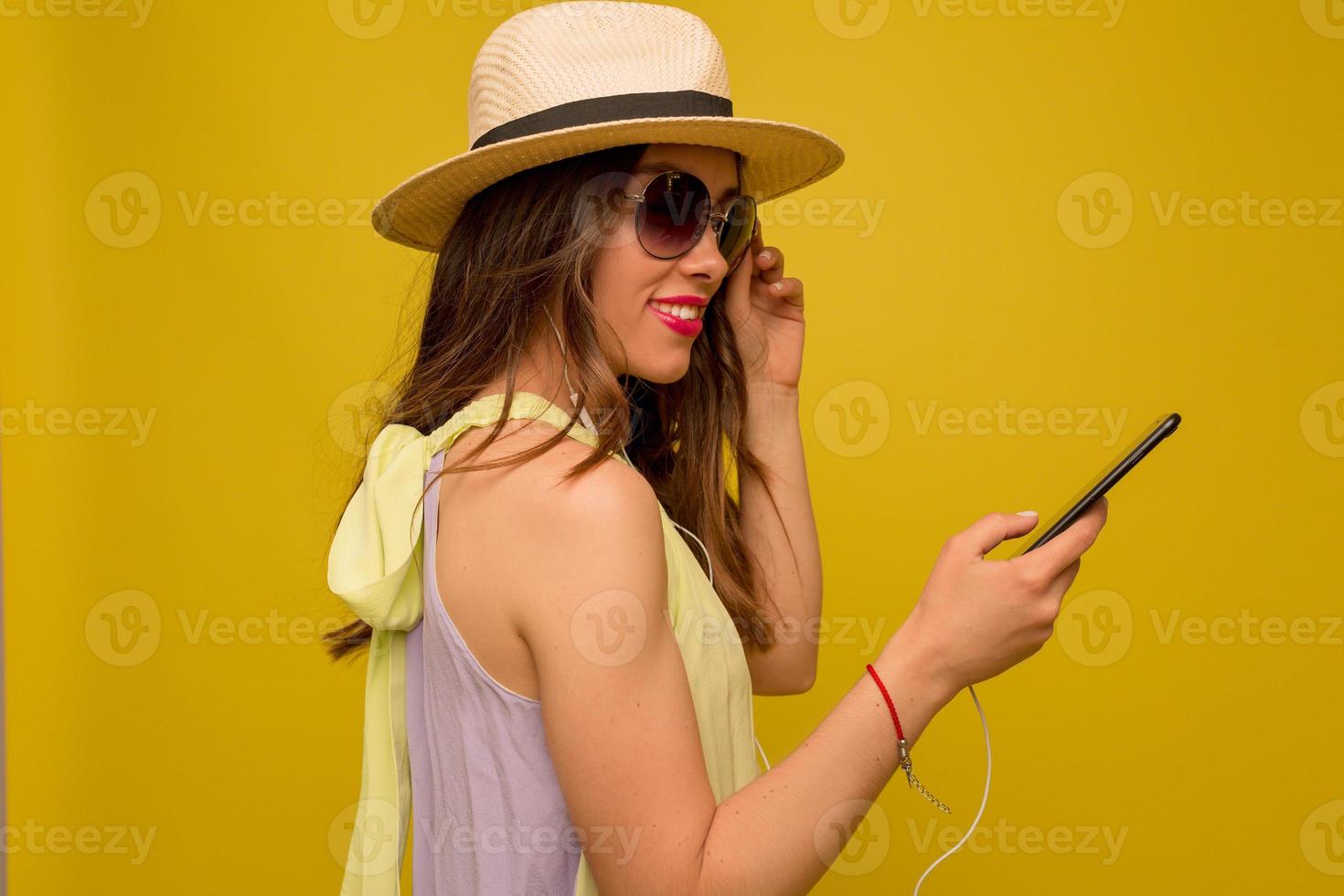 mulher morena bem-humorada com vestido de verão brilhante e chapéu usando smartphone em fundo amarelo. foto