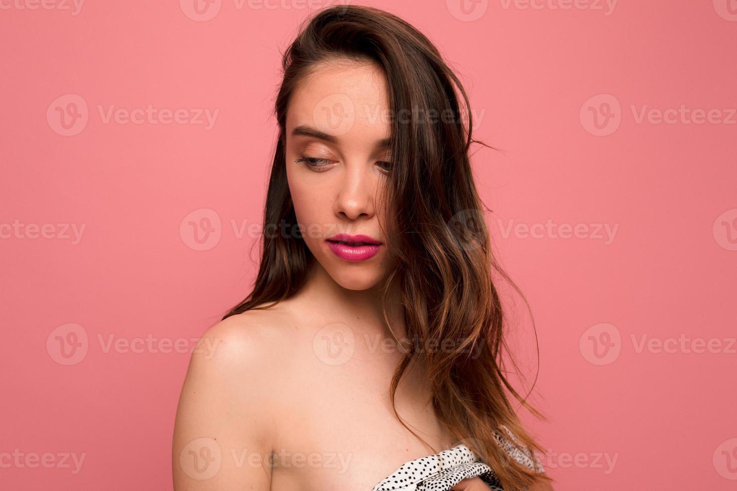 concurso menina elegante com cabelos castanhos ondulados e lábios cor de rosa está olhando para baixo sobre fundo rosa. retrato de senhora encantadora está vestida de vestido com ombros nus e posando em fundo isolado foto