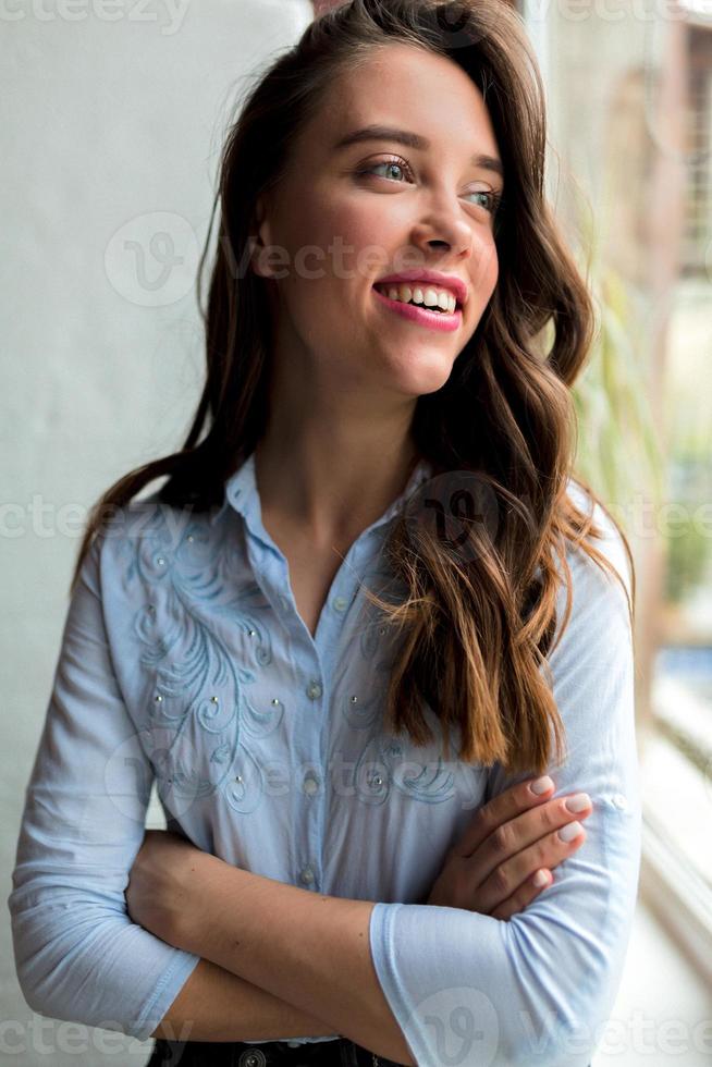 retrato interior de menina bonita atraente com cabelo escuro longo ondulado está vestindo camisa azul posando perto da janela com um sorriso feliz foto