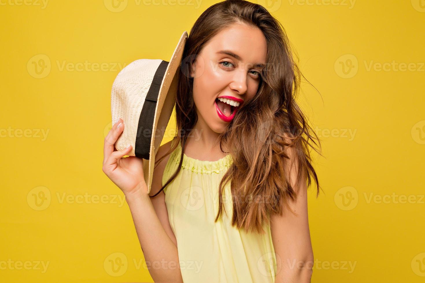 incrível mulher de cabelos compridos vestido de verão brilhante posando com emoções expressivas, ela abriu o moush e segura o chapéu perto do rosto em fundo amarelo foto
