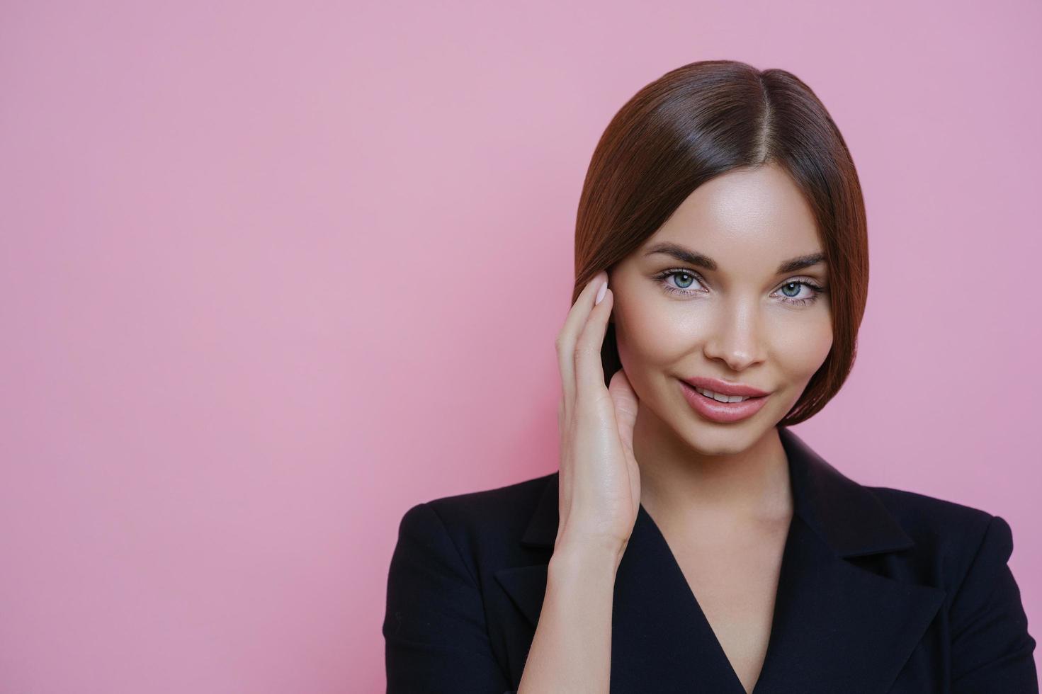 foto de linda empresária europeia com pele saudável, toca o rosto suavemente, retorna do salão spa, usa roupa preta formal, olha com confiança para a câmera, isolada no fundo rosa