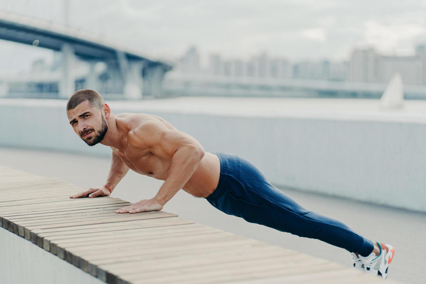 tiro de corpo inteiro de homem musculoso desportivo no sportswear faz push up exercício concentrou poses de expressão séria ao ar livre. auto determinado esportista barbudo prancha por ter braços fortes foto