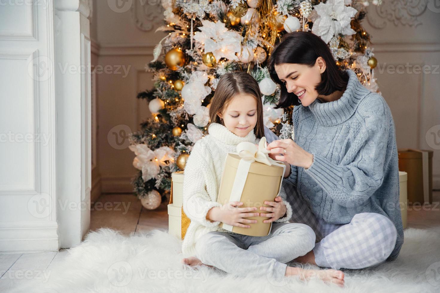 retrato de menina bonita e linda mãe feminina sentam juntos no tapete quente, segure a caixa de presente, aproveite a árvore decorada do ano novo. família passar tempo juntos. conceito de celebração e feriados foto