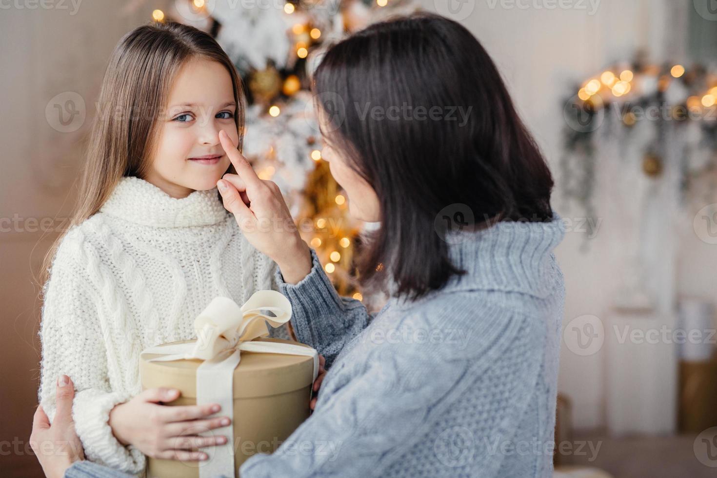 retrato de adorável criança e mãe, toca seu narizinho, dá caixa de presente embrulhada, prepara uma pequena surpresa para a filha, dá presente perto da árvore decorada de ano novo. natal e ano novo foto