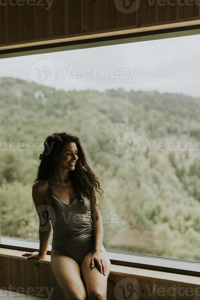 jovem no centro de spa de bem-estar, sentado ao lado da enorme janela larga, apreciando a vista da floresta verde foto