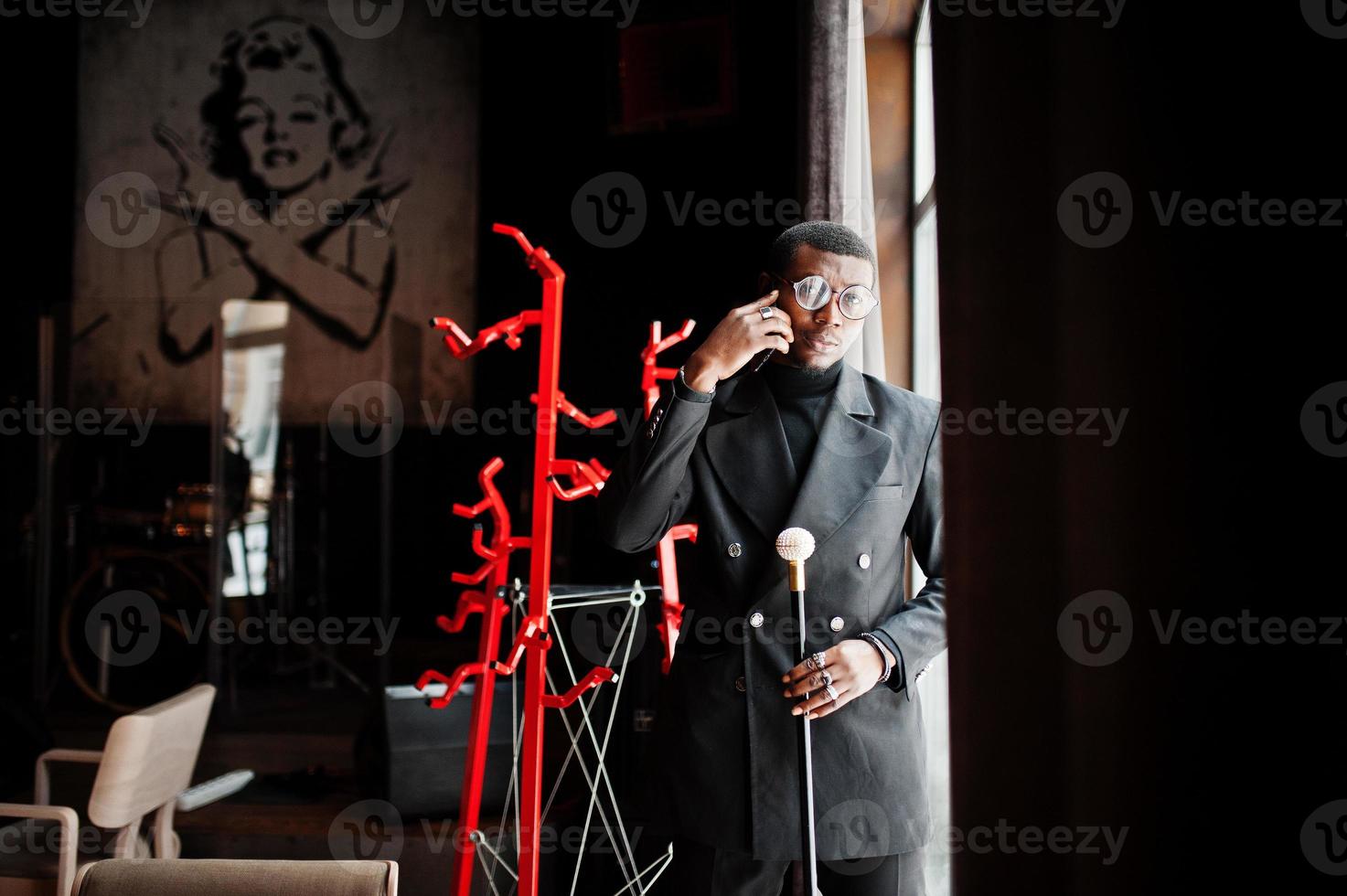 elegante cavalheiro americano africano elegante jaqueta preta e óculos, segurando a bengala retrô como balão de cana ou bengala com alça de bola de diamante dourado e falando no celular. foto