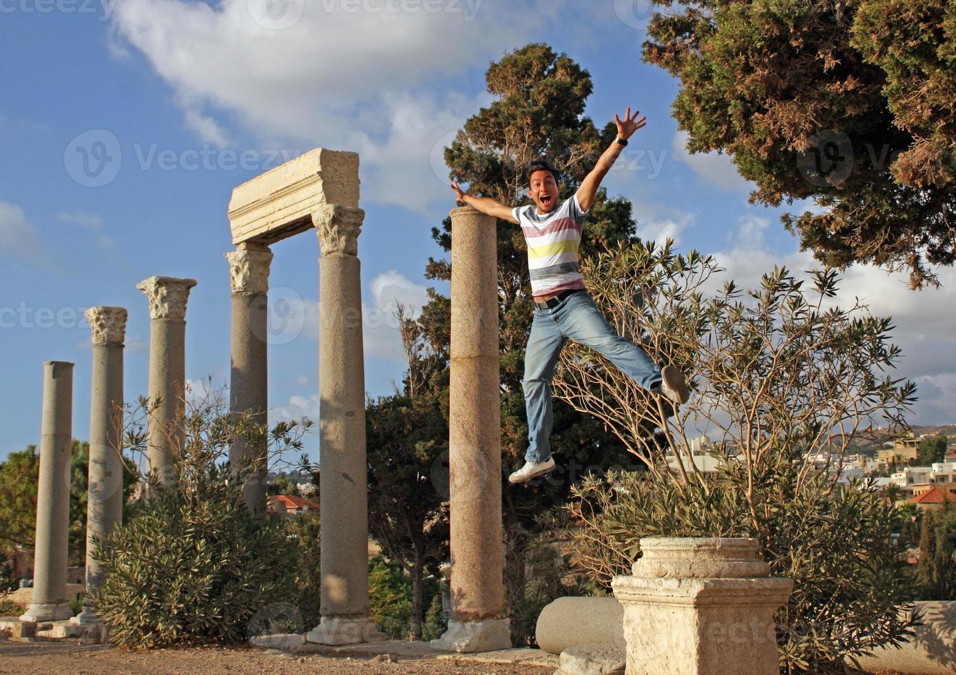 jovem com cara animada pulando com os braços levantados de uma coluna em byblos, líbano. foto