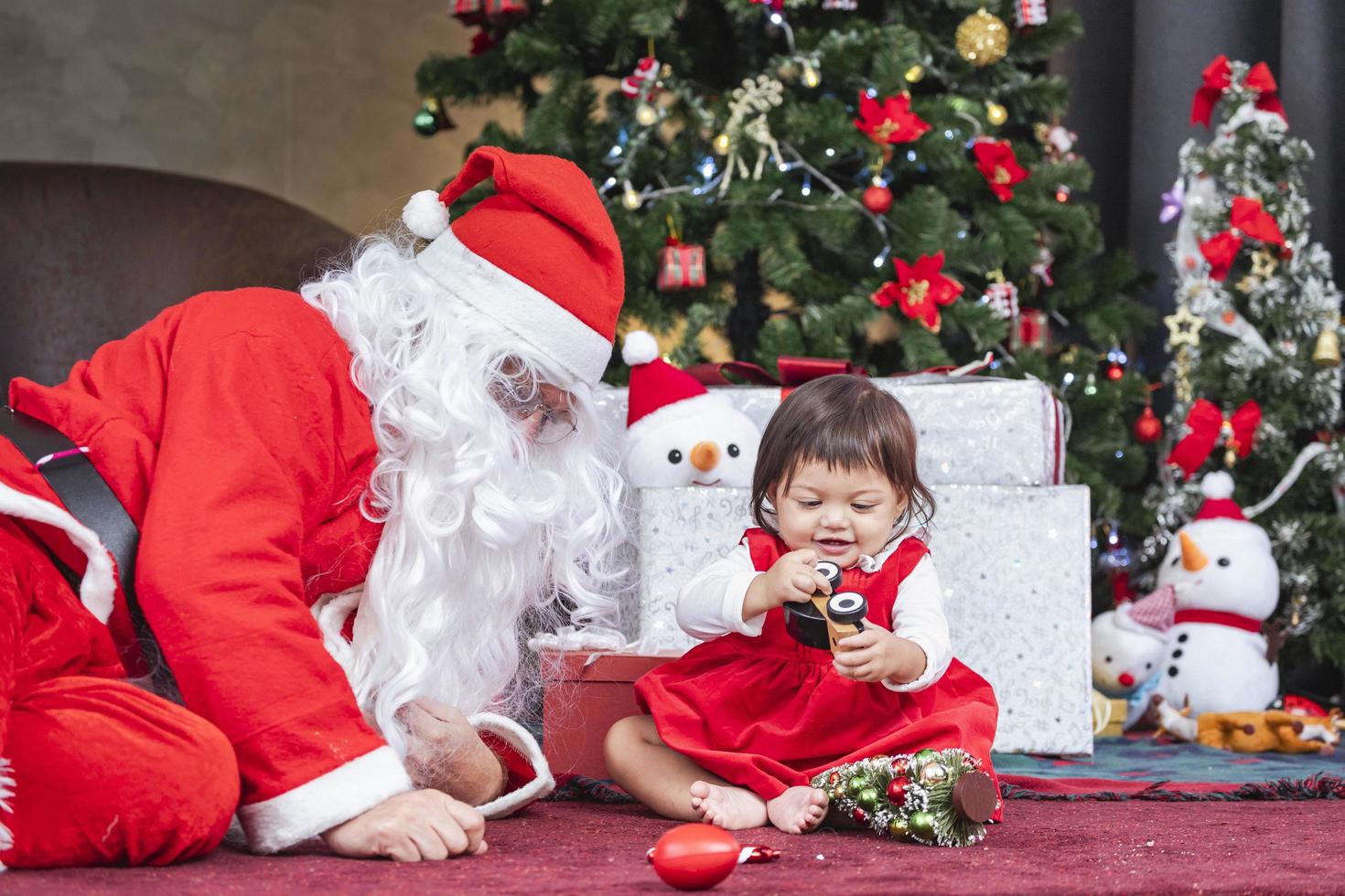 bebê criança está se divertindo brincando com brinquedos enquanto papai noel está preparando o presente e sentado atrás da árvore de natal para o conceito de celebração da temporada foto