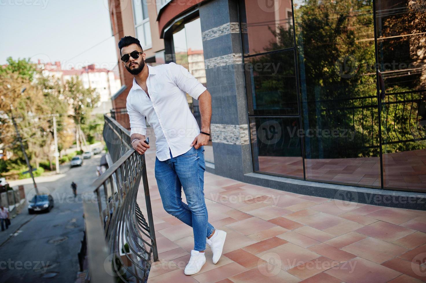 elegante modelo de homem árabe alto na camisa branca, jeans e óculos de sol posou na rua da cidade. barba cara árabe atraente contra edifício moderno. foto