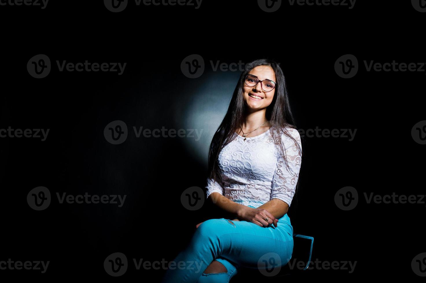 retrato de uma jovem atraente no top branco e calça azul sentado posando com os óculos no escuro. foto