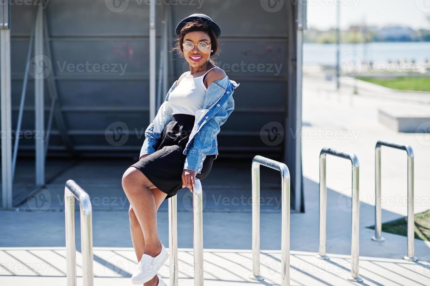 elegante modelo afro-americano de chapéu de óculos, jaqueta jeans e saia preta posou ao ar livre. foto