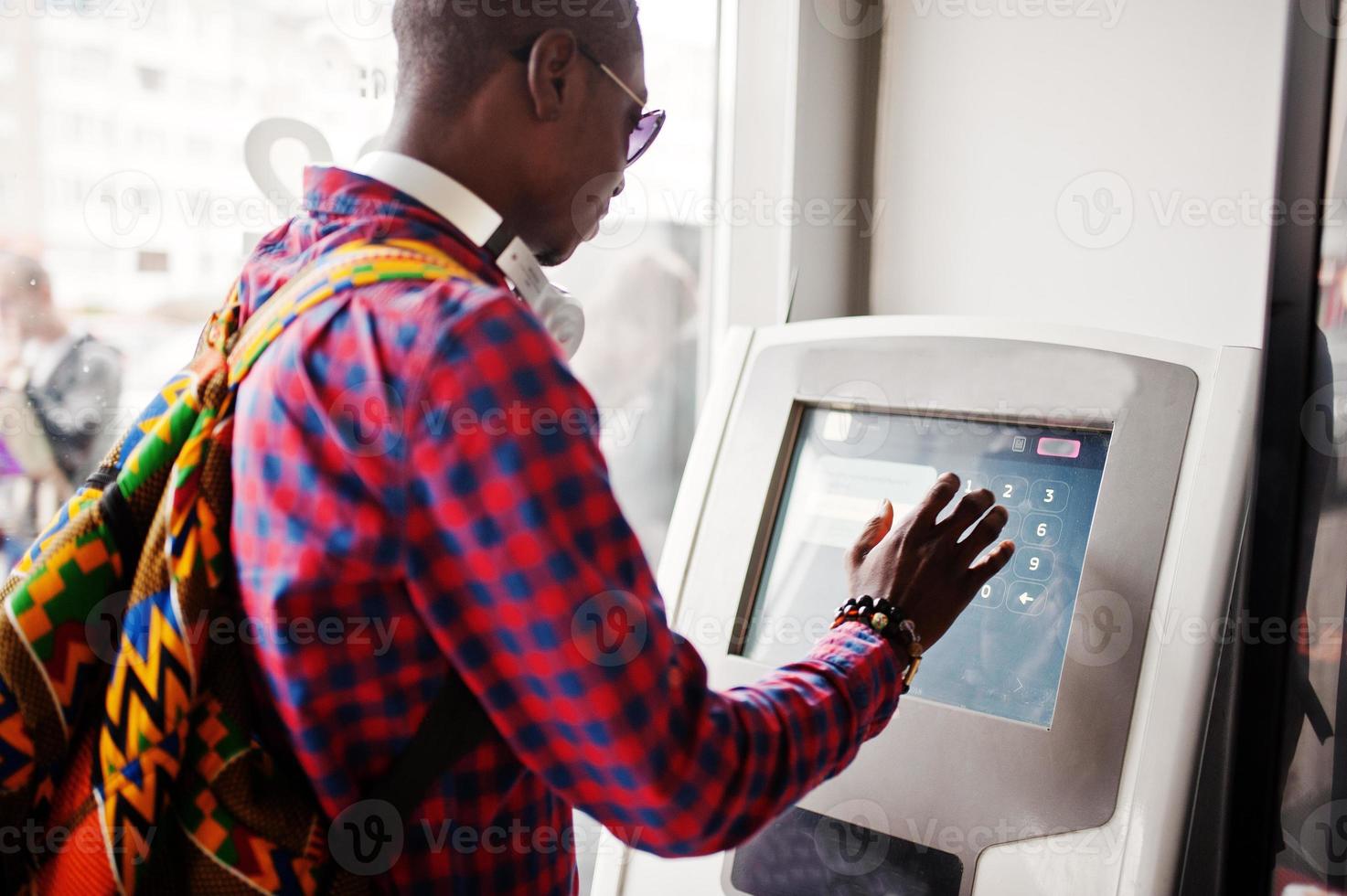 homem afro-americano de camisa quadriculada com mochila segurando a carteira na mão contra o atm. viajante negro. foto