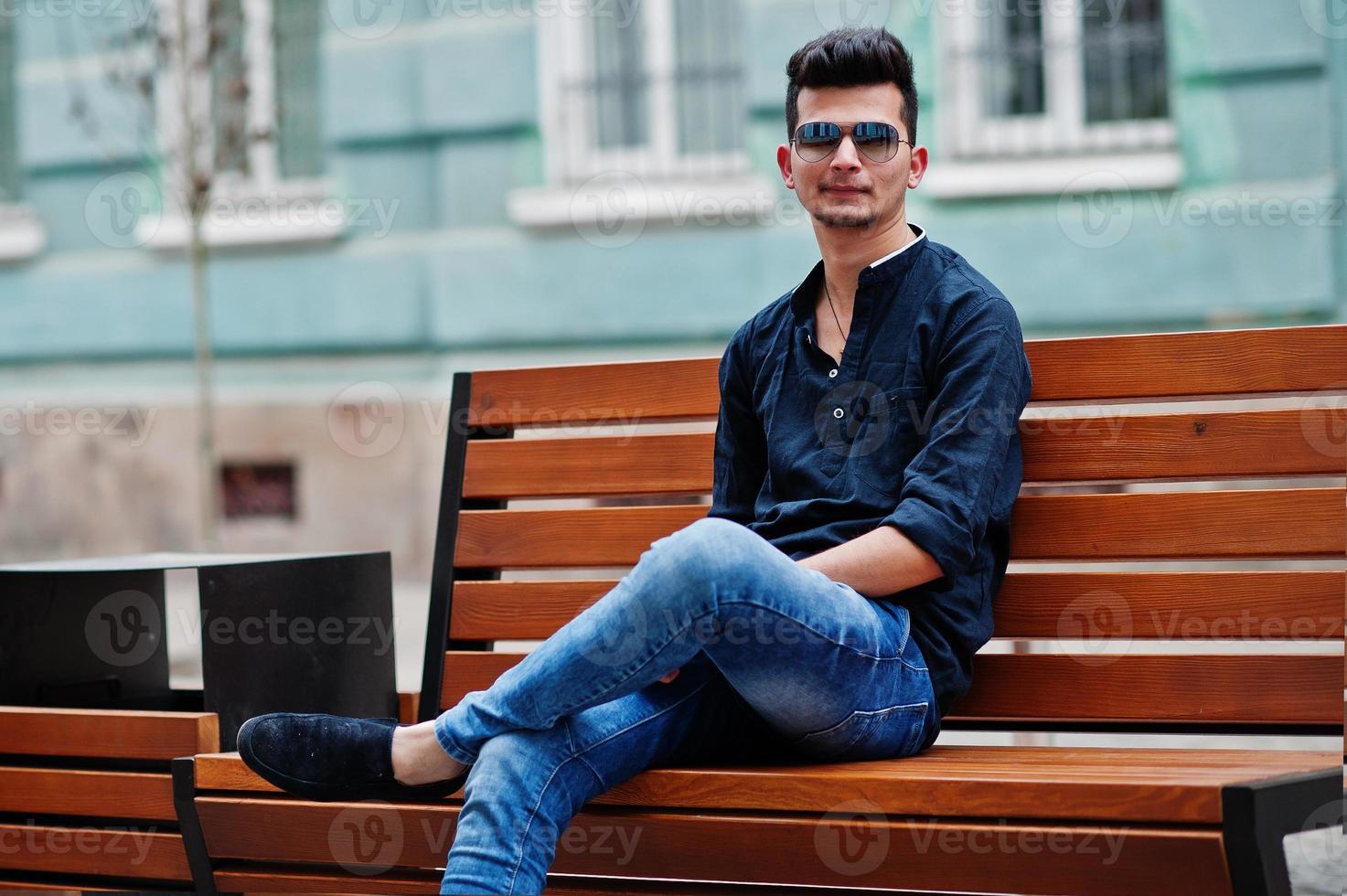 homem elegante modelo indiano em roupas casuais, camisa preta e óculos de sol posou ao ar livre na rua da índia e sentado no banco. foto